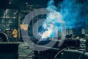 Welder in protective mask works in metallurgical factory workshop, sparks and smoke from welding flares, industrial