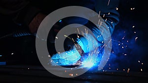 The welder works in a mask in slow motion. Sparks fly in different directions. Blue color glow welding. Work with steel