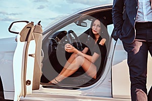 Well-dressed beautiful woman sitting in the front seat and man dressed in suit standing near the car outdoors.