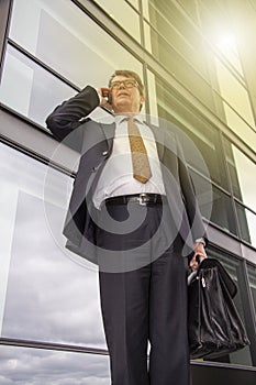 Well dressed businessman with briefcase standing talking on the phone in front of modern glass building  with sun reflection. Pers