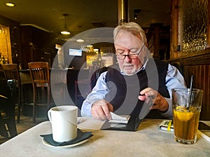 Well dressed senior man in resturant by bar putting signed credit card receipt back into folder with coffee an iced tea sitting on