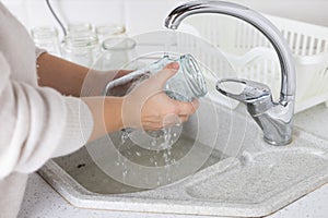 Well in the kitchen, a woman washes glass jars in the sink to sort them for recycling. The woman is responsible for