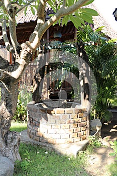 Well in tropical garden with brick paneling. Strong boat rope suspended on a pulley for pulling a pail with water. Wooden bungalow