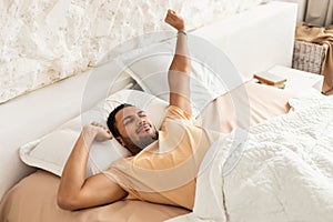 Wellslept Middle Eastern Man Awakening Stretching Arms Lying In Bedroom