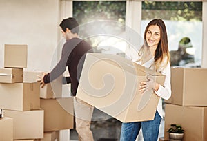 Were moving house, box by box. Portrait of an attractive young woman carrying a box with her husbands in the background.