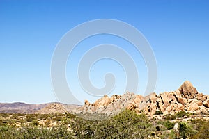 Western desert landscape with green brush in the foreground and rock formations and mountains in the background