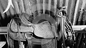 A western saddle and tack sitting on a rail in a barn in black and white photo