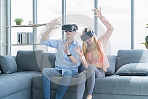 A western young couple wearing VR or virtual reality glasses, headsets sitting and playing a video game on sofa in front of TV at