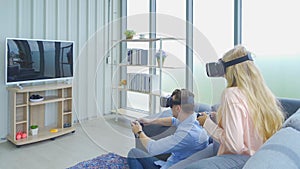 A western young couple wearing VR or virtual reality glasses, headsets sitting and playing a video game on sofa in front of TV at