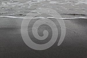 Wet black volcanic sand with sea wave with foam as line on seashore, texture, background.