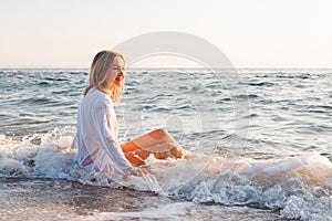 Wet blonde girl in white shirt  sitting in the water on the sea beach and looks at the   the dawn of the sun. Concept: summer