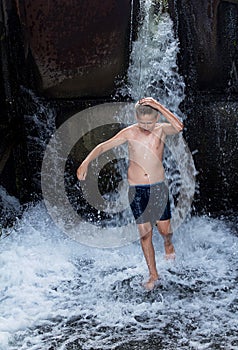 Wet boy in shorts, running in the water, next to a waterfall