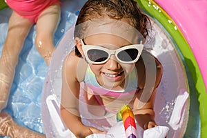 A wet child in a swimsuit and sunglasses is sitting in an inflatable pool