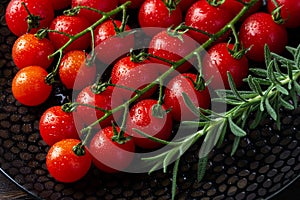 Wet ripe red cherry tomatoes on the vine and sprig of fresh rosemary on a black plate. Macro. Water drops on a small round