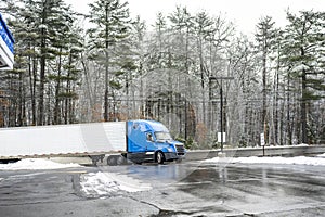 Wet shiny blue big rig semi truck with refrigerator semi trailer turning on the truck stop entrance with snow and ice slippery