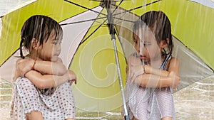 Wet siblings little girl sitting and hugging knees under an umbrella on rainy day at yard with sad emotion.