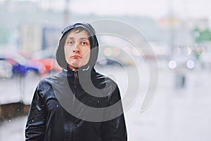 Wet young man in raincoat with unhappy face walks along the street during heavy rain, close-up. Autumn bad stormy weather concept