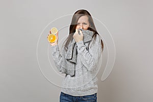 Whacked young woman in gray sweater covering mouth with scarf, sneezing or coughing, hold lemon, orange isolated on grey photo