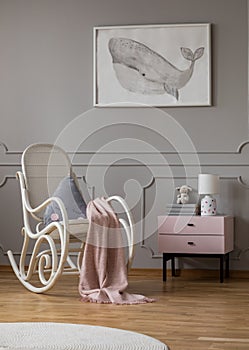 Whale on poster in baby room with white rocking chair and pastel pink nightstand with books and lamp