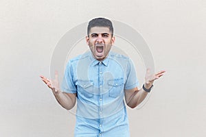 What do you need? Portrait of angry young bearded man in blue shirt standing with raised arms and looking at camera, asking and