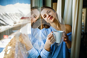 Portrait of a joyful young woman enjoying a cup of coffee at home. Smiling beautiful girl drinks hot tea in winter.