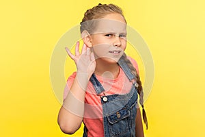 What? I can`t hear! Portrait of curious attentive little girl in denim overalls holding hand near ear