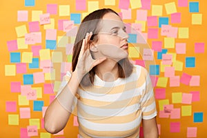 What? I can`t hear you. Confused woman standing isolated over yellow background with memo cards, keeping hand near ear to listen