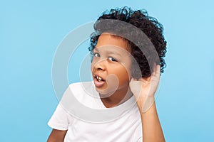 What? I can`t hear you. Portrait of attentive curious little boy with curly hair holding hand near ear and listening carefully photo