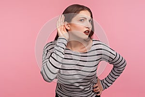 What? I can`t hear you! Portrait of confused woman in striped sweatshirt keeping hand near ear to listen better