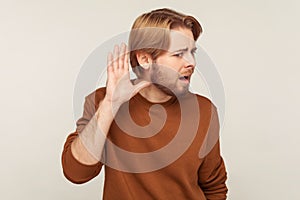 What? I don`t hear you! Portrait of bearded man in sweatshirt standing with hand near ear and listening to whisper photo