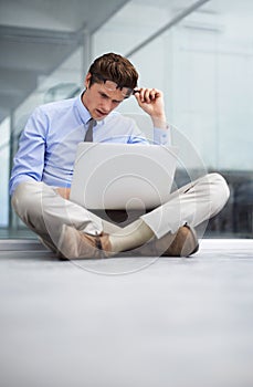 What This makes no sense. A young businessman sitting on the floor of his empty office working on a laptop.