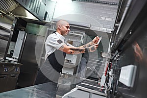 What is next... Handsome bald male chef with tattoos on his arms looking at order list in a restaurant kitchen.