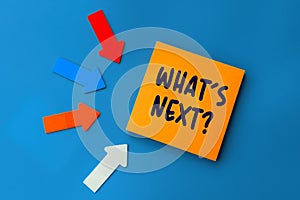 What`s Next - concept of text on sticky note. Orange square sticky note and colorful arrows on blue background