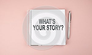 WHAT`S YOUR STORY text on notebook with pen on the pink background