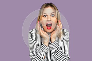 What a surprise! Excited blond woman in striped blouse standing with wide open mouth and raising hands in amazement