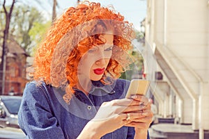 What??? Unhappy frustrated angry woman with red curly hair looking to mobile phone, reading something, standing outside with city