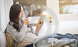 Whats healthy for you is healthy for your baby. an attractive young pregnant woman eating fruit and reading a book while