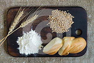 Wheat ears, grains, flour and sliced bread on a kitchen board on a sacking background