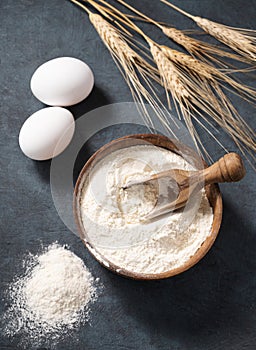 Wheat flour in a wooden bowl with a scoop on a dark blue background with eggs and dry branches wheat. Organic ingredients for