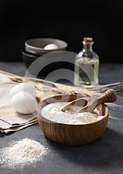 Wheat flour in a wooden bowl with a scoop on a dark blue background with eggs, oil and dry branches wheat. Organic ingredients for