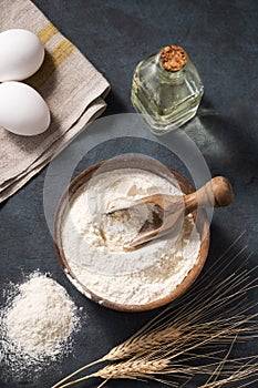 Wheat flour in a wooden bowl with a scoop on a dark blue background with eggs, oil and dry branches wheat. Organic ingredients for