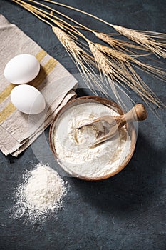 Wheat flour in a wooden bowl with a scoop and eggs on a dark background. Natural organic flour for home baking