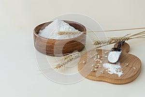 wheat flour in a wooden bowl and in a spoon on a board, near ears of wheat and some grains on a light background.