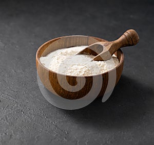 Wheat or oat flour in a wooden bowl with scoop on a dark background. Organic baking ingredients. Concept of homemade and healthy