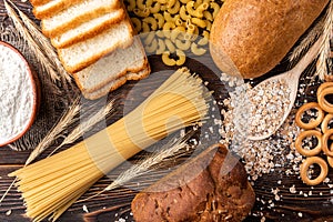 Wheat products on dark wooden background. Bread, spaghetti, wheat flour and flakes, loaf and dryings.