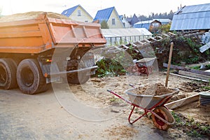 wheelbarrow with sand and shovel in foreground and dump truck preparing to dump sand on ground in background
