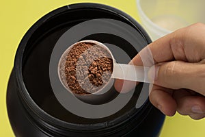 Whey Protein. Point of view of hand holding measuring scoop. Chocolate flavour. Color background: yellow