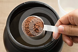 Whey Protein. Point of view of hand holding measuring scoop. Chocolate flavour. Wooden background