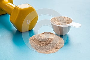 Whey protein powder in plastic measuring scoop near dumbbells on blue background