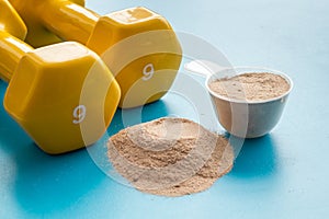 Whey protein powder in plastic measuring scoop near dumbbells on blue background